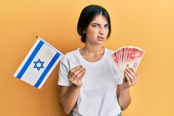 Young caucasian woman holding israel flag and shekels banknotes skeptic and nervous, frowning upset...