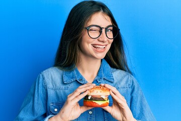 Young caucasian woman eating a tasty classic burger winking looking at the camera with sexy...
