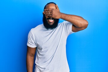 Young african american man wearing casual white tshirt smiling and laughing with hand on face covering eyes for surprise. blind concept.