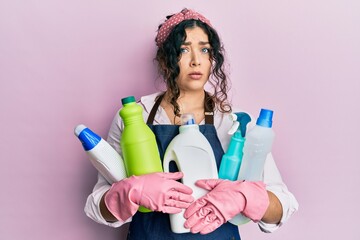 Young brunette woman with curly hair wearing cleaner apron holding cleaning products in shock face,...