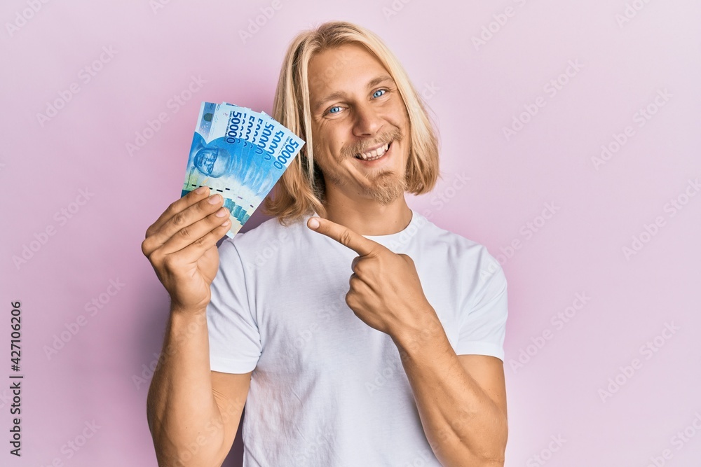 Sticker caucasian young man with long hair holding 50000 indonesian rupiah smiling happy pointing with hand 