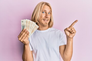 Caucasian young man with long hair holding 100 danish krone banknotes smiling happy pointing with hand and finger to the side
