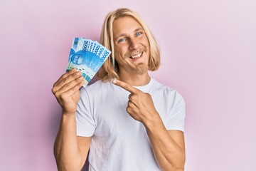 Caucasian young man with long hair holding 50000 indonesian rupiah smiling happy pointing with hand and finger