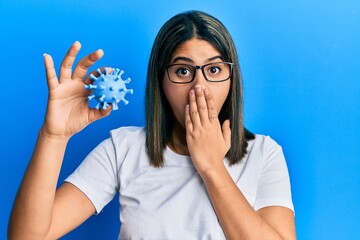 Young latin woman holding virus toy covering mouth with hand, shocked and afraid for mistake....
