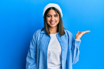 Young latin woman wearing cute wool cap smiling cheerful presenting and pointing with palm of hand looking at the camera.