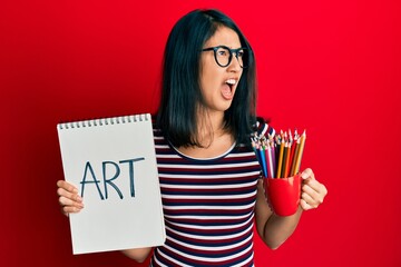 Beautiful asian young woman holding art notebook and colored pencils angry and mad screaming frustrated and furious, shouting with anger. rage and aggressive concept.