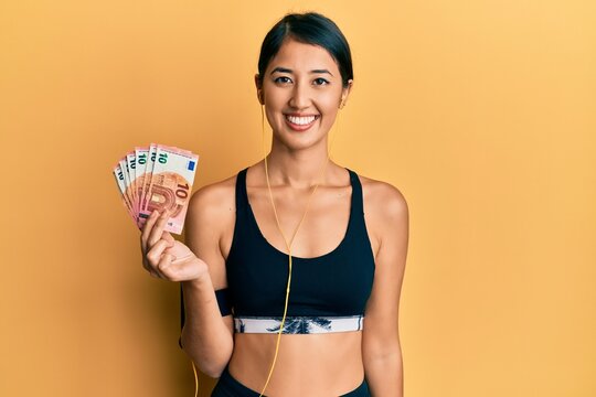 Beautiful asian young sport woman holding bunch of 50 euro banknotes looking positive and happy standing and smiling with a confident smile showing teeth