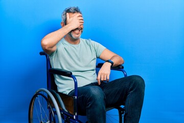 Fototapeta premium Handsome middle age man with grey hair sitting on wheelchair smiling and laughing with hand on face covering eyes for surprise. blind concept.