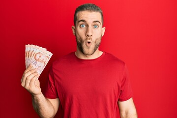 Young caucasian man holding 10 singapore dollars banknotes scared and amazed with open mouth for surprise, disbelief face