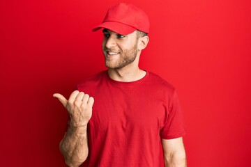 Young caucasian man wearing delivery uniform and cap smiling with happy face looking and pointing to the side with thumb up.