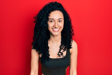 Young hispanic woman with curly hair wearing casual style with sleeveless shirt looking positive and happy standing and smiling with a confident smile showing teeth