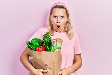 Beautiful caucasian blonde woman holding paper bag with groceries scared and amazed with open mouth for surprise, disbelief face