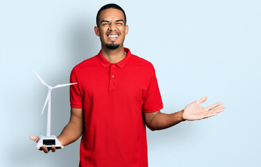 Young african american man holding solar windmill for renewable electricity celebrating achievement with happy smile and winner expression with raised hand