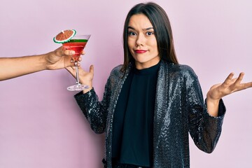 Young girl wearing sexy party jacket and hispanic arm offering cocktail clueless and confused with open arms, no idea and doubtful face.