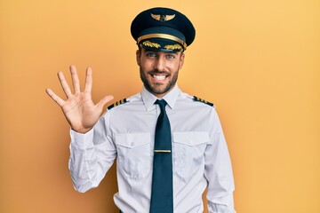 Handsome hispanic man wearing airplane pilot uniform showing and pointing up with fingers number five while smiling confident and happy. © Krakenimages.com