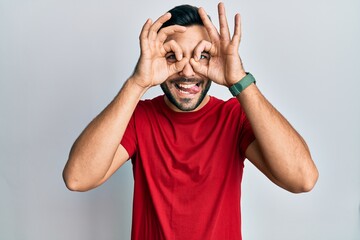 Young hispanic man wearing casual clothes doing ok gesture like binoculars sticking tongue out, eyes looking through fingers. crazy expression.