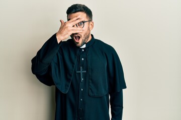 Obraz premium Young hispanic man wearing priest uniform standing over white background peeking in shock covering face and eyes with hand, looking through fingers with embarrassed expression.