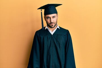 Young hispanic man wearing graduation cap and ceremony robe skeptic and nervous, frowning upset...