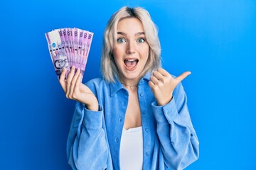 Young blonde girl holding philippine peso banknotes pointing thumb up to the side smiling happy with open mouth