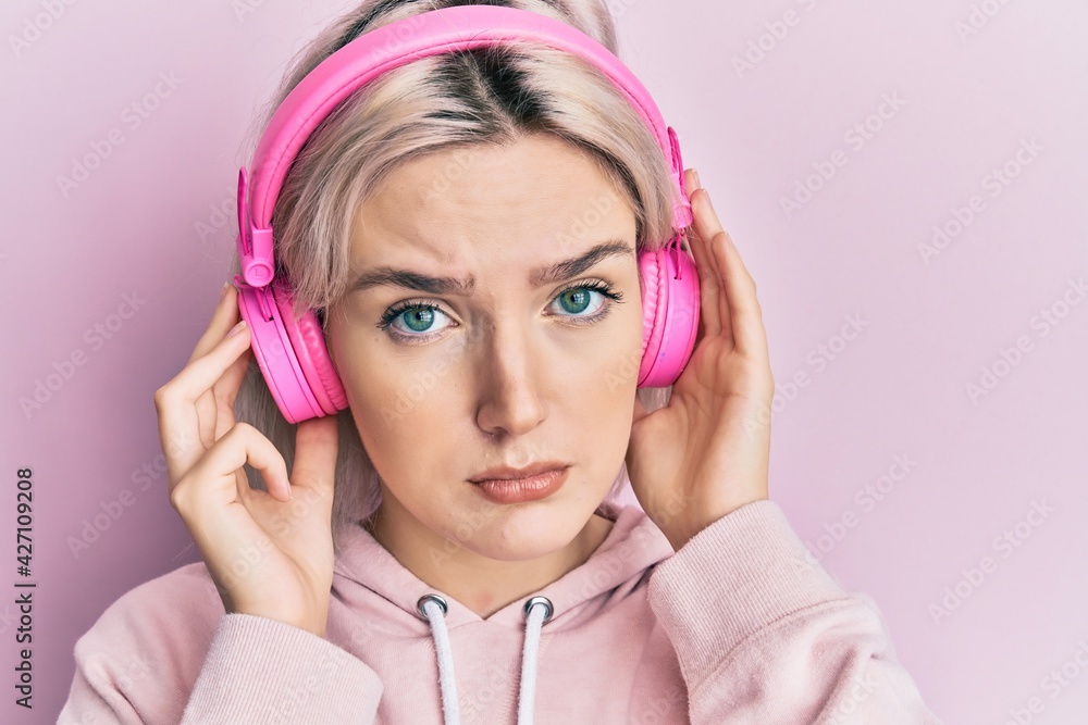 Poster young blonde girl listening to music using headphones skeptic and nervous, frowning upset because of