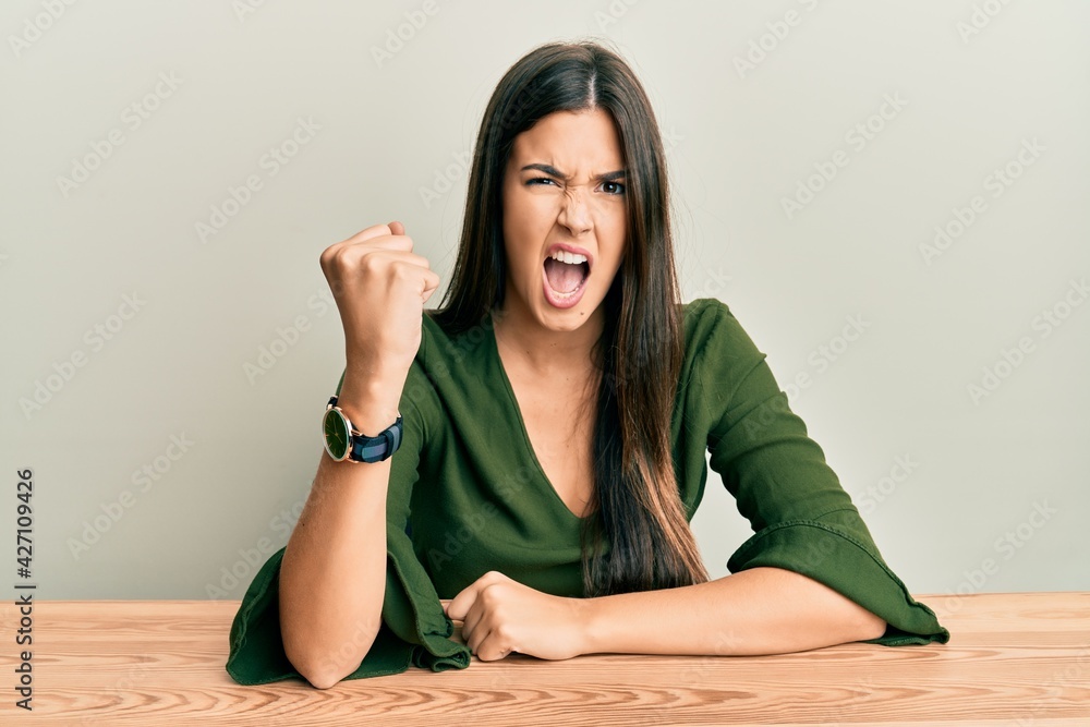 Poster Young brunette woman wearing casual clothes sitting on the table angry and mad raising fist frustrated and furious while shouting with anger. rage and aggressive concept.