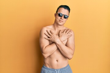 Fototapeta premium Handsome young man wearing swimsuit and sunglasses hugging oneself happy and positive, smiling confident. self love and self care