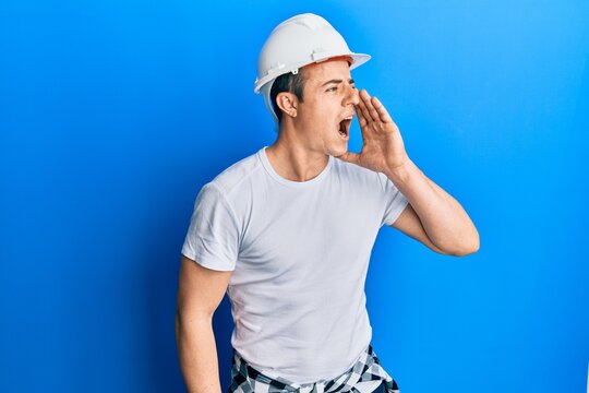 Handsome young man wearing builder uniform and hardhat shouting and screaming loud to side with hand on mouth. communication concept.