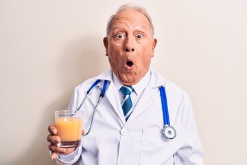 Senior grey-haired doctor man wearing stethoscope drinking glass of healthy orange juice scared and...
