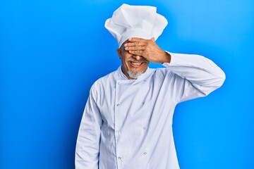 Middle age grey-haired man wearing professional cook uniform and hat smiling and laughing with hand on face covering eyes for surprise. blind concept.