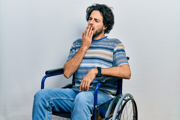 Handsome hispanic man sitting on wheelchair bored yawning tired covering mouth with hand. restless...