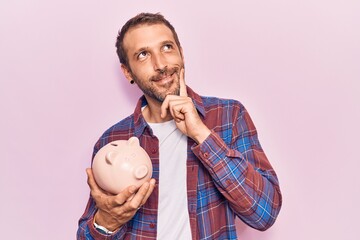 Young handsome man holding piggy bank serious face thinking about question with hand on chin,...