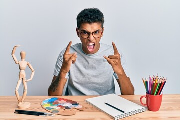 Naklejka premium Young handsome african american man painter sitting palette and art manikin shouting with crazy expression doing rock symbol with hands up. music star. heavy concept.
