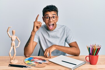 Young handsome african american man painter sitting palette and art manikin pointing finger up with successful idea. exited and happy. number one.