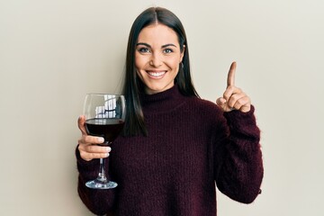 Young brunette woman drinking a glass of red wine smiling with an idea or question pointing finger...