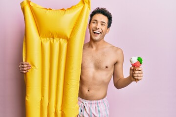 Obraz premium Young handsome man wearing swimsuit holding summer mattress float and icecream smiling and laughing hard out loud because funny crazy joke.