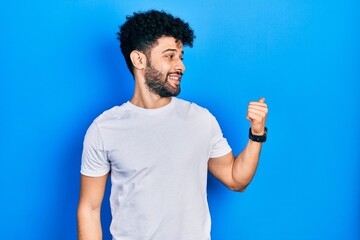 Young arab man with beard wearing casual white t shirt smiling with happy face looking and pointing to the side with thumb up.