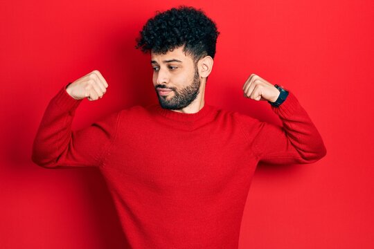 Young arab man with beard wearing casual red sweater showing arms muscles smiling proud. fitness concept.
