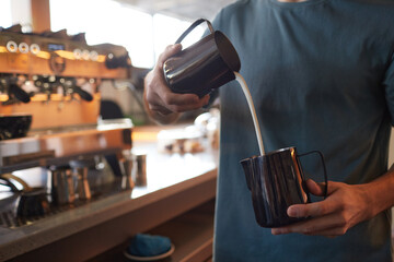 Cropped portrait of young barista pouring cream in latte while making fresh coffee in cafe or coffee shop, copy space