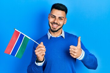Young hispanic man with beard holding gambia flag smiling happy and positive, thumb up doing excellent and approval sign