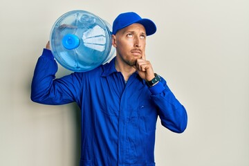 Bald courier man with beard holding a gallon bottle of water for delivery serious face thinking about question with hand on chin, thoughtful about confusing idea