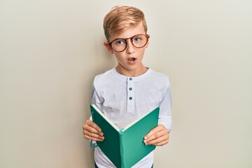 Little caucasian boy kid reading a book wearing glasses in shock face, looking skeptical and sarcastic, surprised with open mouth
