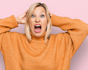 Middle age caucasian woman wearing casual winter sweater crazy and scared with hands on head,...