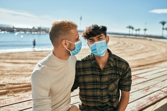 Young gay couple wearing medical mask sitting on the bench at the beach.