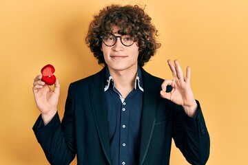 Handsome young man holding engagement ring for proposal doing ok sign with fingers, smiling...