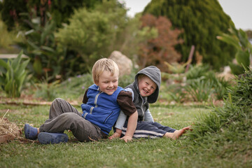 Beautiful blonde brothers sitting back to back on grass in cute garden with happy smiles
