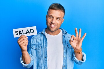 Handsome muscle man holding salad word on paper doing ok sign with fingers, smiling friendly gesturing excellent symbol