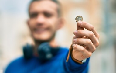 Young hispanic man using headphones holding 1 euro coin at the city