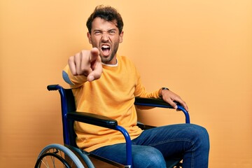 Handsome man with beard sitting on wheelchair pointing displeased and frustrated to the camera, angry and furious with you