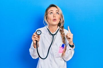 Beautiful young blonde doctor woman holding stethoscope amazed and surprised looking up and pointing with fingers and raised arms.