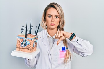 Beautiful young blonde woman holding model of anatomical skin and hair with angry face, negative...